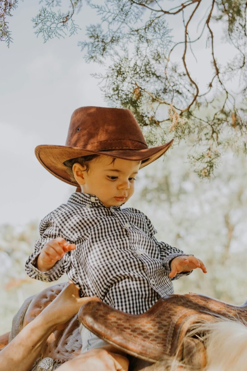 Charming baby in cowboy attire sitting on a saddle surrounded by nature.